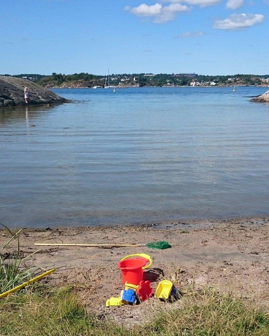 Paradisbukta: sandstrand med bøtte og spade, rolig sjø og svaberg på hver side.